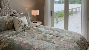 A bedroom with a large bed, a nightstand, a lamp, and a view of a body of water through a sliding glass door. - Rivendock Rookery II: Waterfront Home w/Dock & Stunning Views (Oriental)