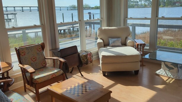 A spacious room with large windows overlooking a body of water and a pier. There are multiple seating options including a sofa, chairs, and a small table. - Rivendock Rookery II: Waterfront Home w/Dock & Stunning Views (Oriental)