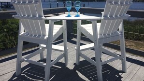 Two white Adirondack chairs with a small table and two blue glasses on it, set on a wooden deck with a view of a body of water and a distant shoreline. - Rivendock Rookery II: Waterfront Home w/Dock & Stunning Views (Oriental)