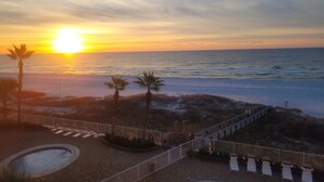 A sunset view from a balcony overlooking a beach with palm trees and a pool.