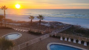 A pool area with a view of the sunset over the ocean, palm trees, and a wooden deck with lounge chairs.
