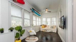 Un salon avec un canapé blanc, une table basse et un téléviseur fixé au mur. De grandes fenêtres sont équipées de stores, un ventilateur de plafond est installé au plafond et un bouée de sauvetage rouge est accrochée au mur.