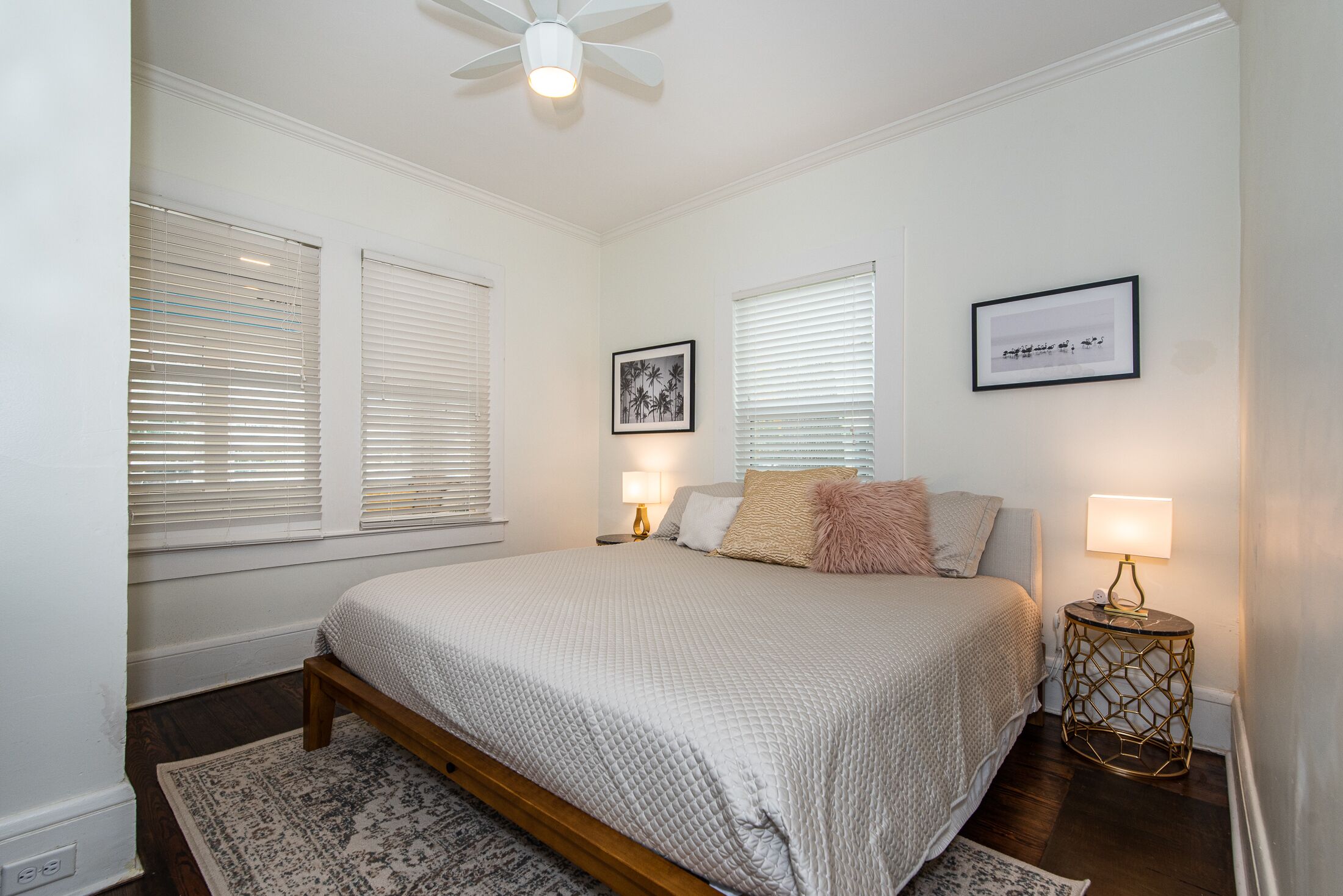 A bedroom with a bed, two bedside lamps, a ceiling fan, and two framed pictures on the wall.