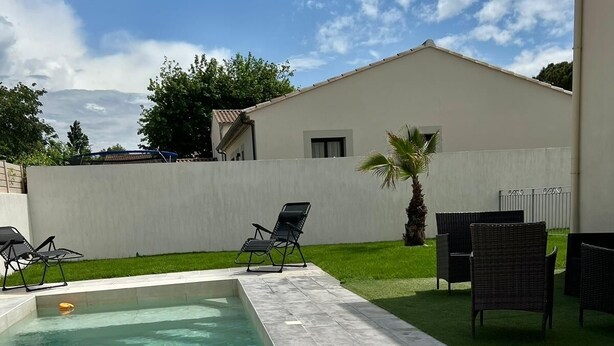 A swimming pool with tiled flooring, surrounded by a grassy area and a white wall. There are lounge chairs and a palm tree. - Villa festival avignon (Avignon)