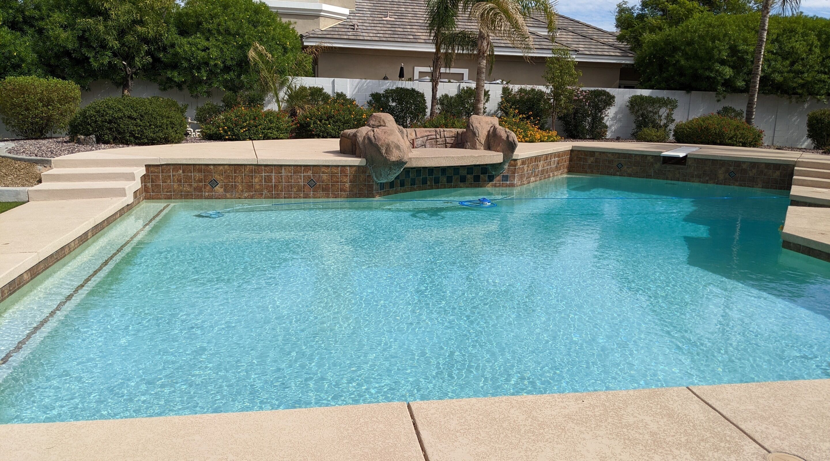 A rectangular swimming pool with clear blue water, surrounded by a tiled deck and a landscaped area with rocks and plants.