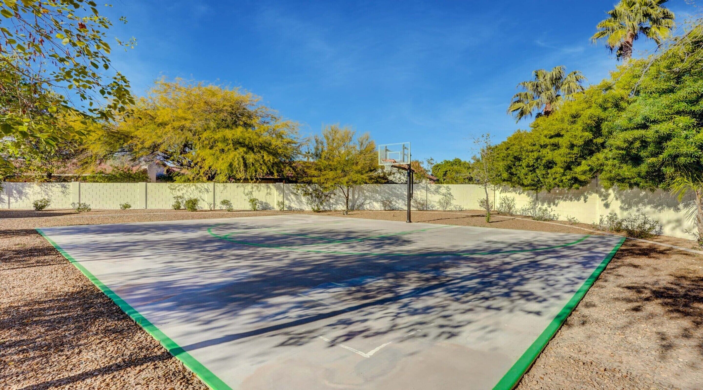 An outdoor basketball court with a green and white boundary, surrounded by trees and a white wall.
