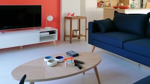 A modern living room with a flat-screen TV, a blue sofa, a coffee table with bowls and a book, and a kitchen in the background.