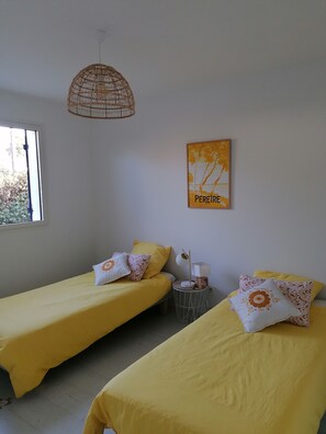 Habitación con dos camas, cada una con ropa de cama amarilla y almohadas decorativas, una mesita de noche pequeña con lámpara, una lámpara colgante de mimbre y un cuadro enmarcado en la pared.