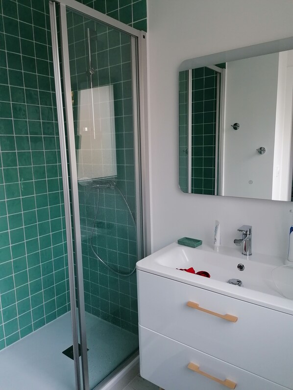 Een badkamer met een douchecabine van glas, een wit wastafelmeubel met een spiegel erboven en groene betegelde wanden.