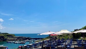 A seaside restaurant with outdoor seating, a rocky coastline, and boats docked in the background.