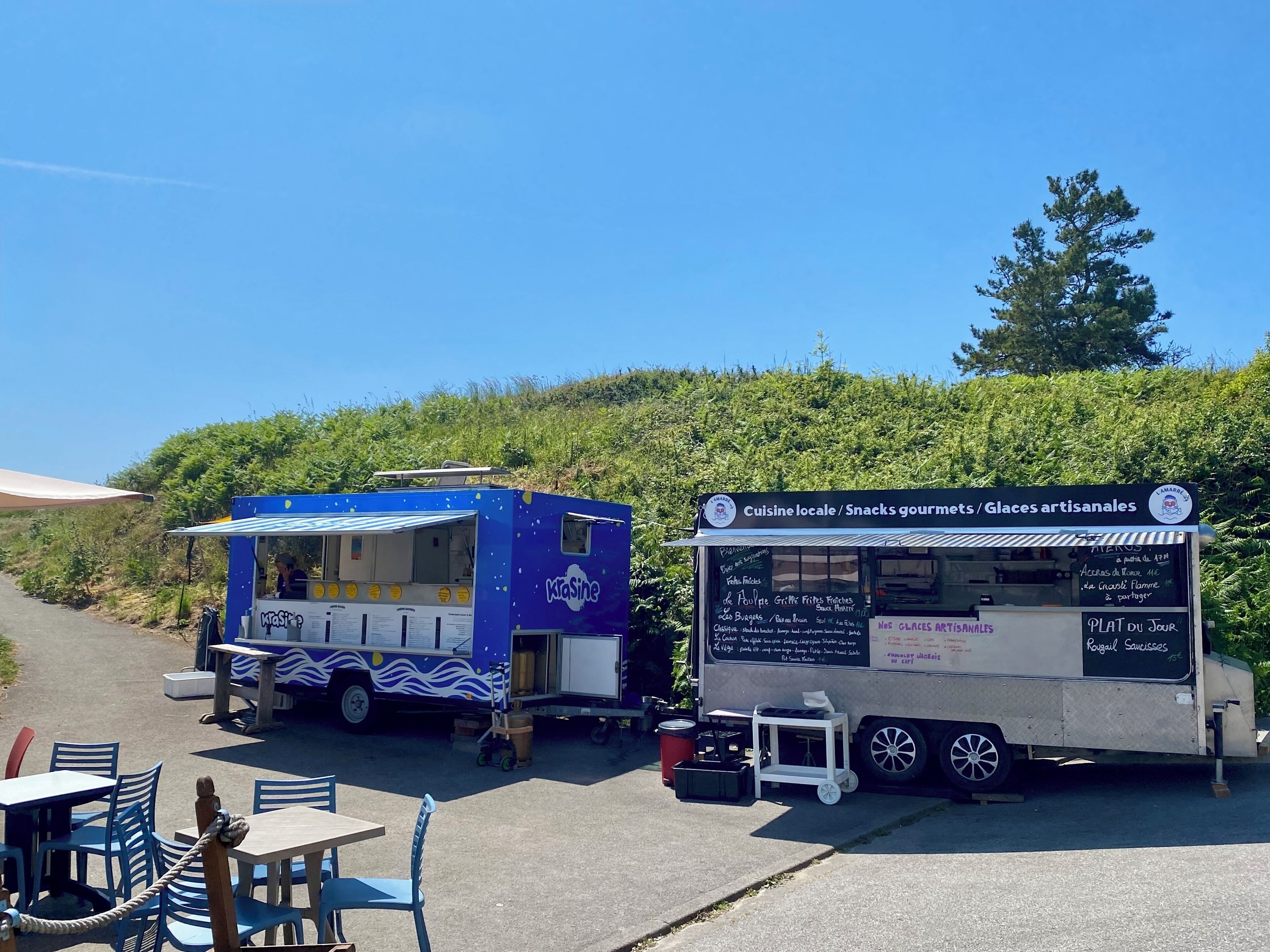 Two food trucks parked on a paved area with tables and chairs nearby.