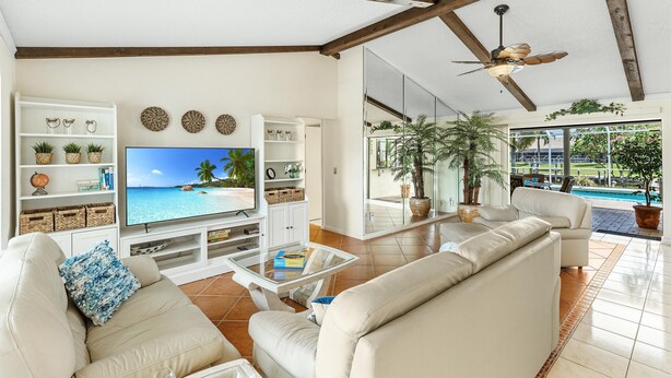Una sala de estar con televisor, sofás blancos, una mesa de centro y vista a una piscina.