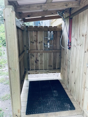 A wooden outdoor shower enclosure with a metal grate floor, a wooden fence, and a metal shelf with bottles.