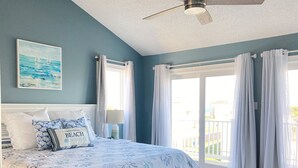 Une chambre à coucher avec un lit, un ventilateur de plafond, une fenêtre avec des rideaux et un tableau au mur.