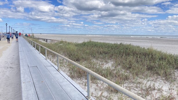 Une promenade en bois bordée d’une rambarde, qui longe une plage et mène au bord de l’eau.