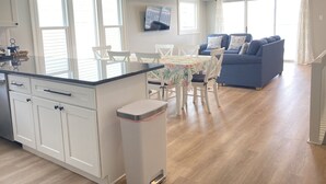 Une cuisine moderne avec des meubles blancs, un îlot central et un coin repas comprenant une table et des chaises. Le salon est doté d’un canapé bleu et d’un téléviseur fixé au mur.