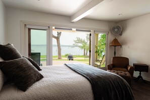 Ein Schlafzimmer mit einem großen Bett, einem Sessel, einer Lampe und Blick auf ein Gewässer durch eine Schiebetür.