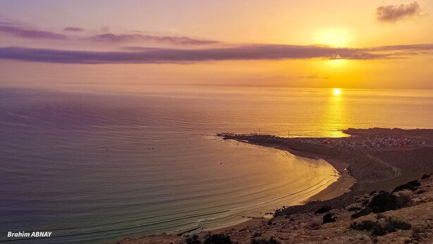 Paisagem costeira com pôr do sol sobre o oceano, uma linha de costa sinuosa e um povoado ao longe.