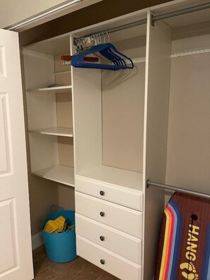 A closet with open doors, featuring a blue hanger, a wooden board with 'HANG' and 'FOOT' text, and a blue basket on the floor.
