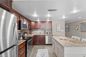 A modern kitchen with stainless steel appliances, wooden cabinets, and a granite countertop. - Garden City Beach Oceanfront Condo 1202 | Rising Tides Properties Rental (Murrells Inlet)