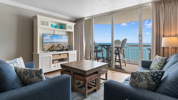 A balcony with a view of the ocean, a dining area with chairs, a table, and a TV on a cabinet. - Garden City Beach Oceanfront Condo 1202 | Rising Tides Properties Rental (Murrells Inlet)