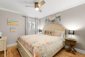 A bedroom with a bed, a ceiling fan, a nightstand with a lamp, and artwork on the walls.