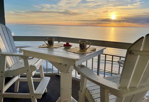 A table with two mugs and a bowl of fruit on a balcony overlooking the sea at sunset.