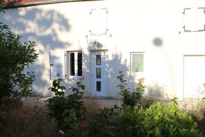 A white building with a door and two windows, surrounded by greenery and a gravel path.