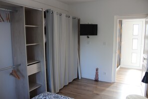 A room with a wooden floor, a bed with a patterned bedspread, a wardrobe with open doors, a flat-screen TV mounted on the wall, and a door leading to another room.