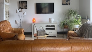 A living room with a leather sofa, a white entertainment unit, a flat-screen TV, a chandelier, and potted plants. - Cottage Philmaries (Sainte-Féréole)