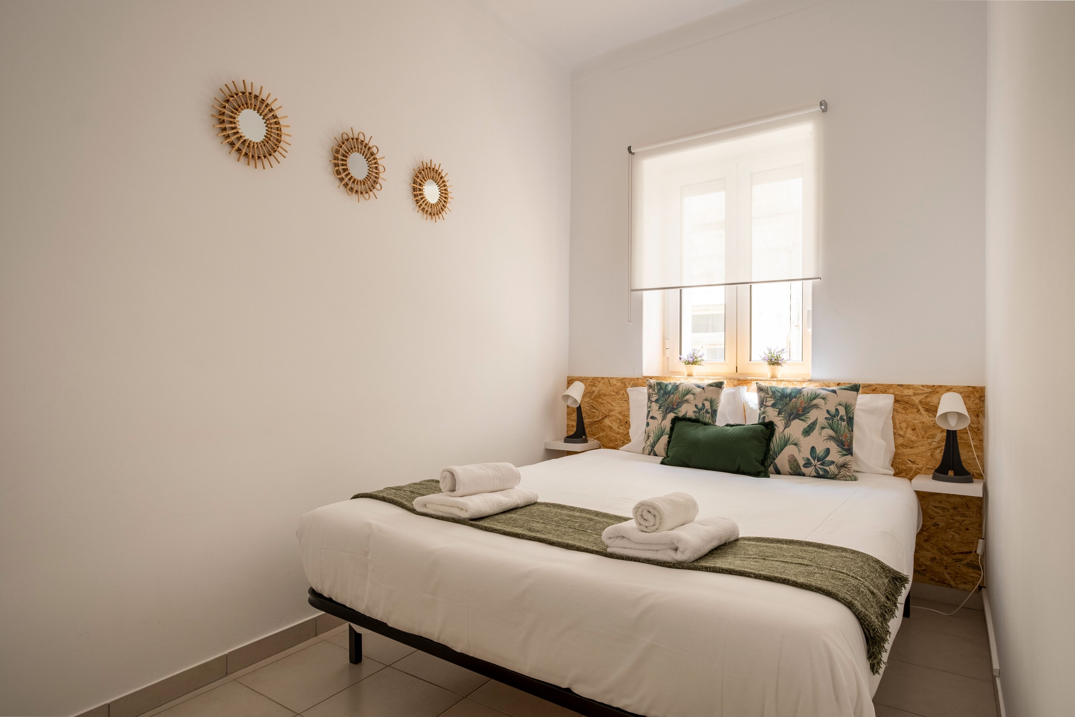 A neatly made bed with white linens and rolled towels, a wooden headboard, and a window with blinds.