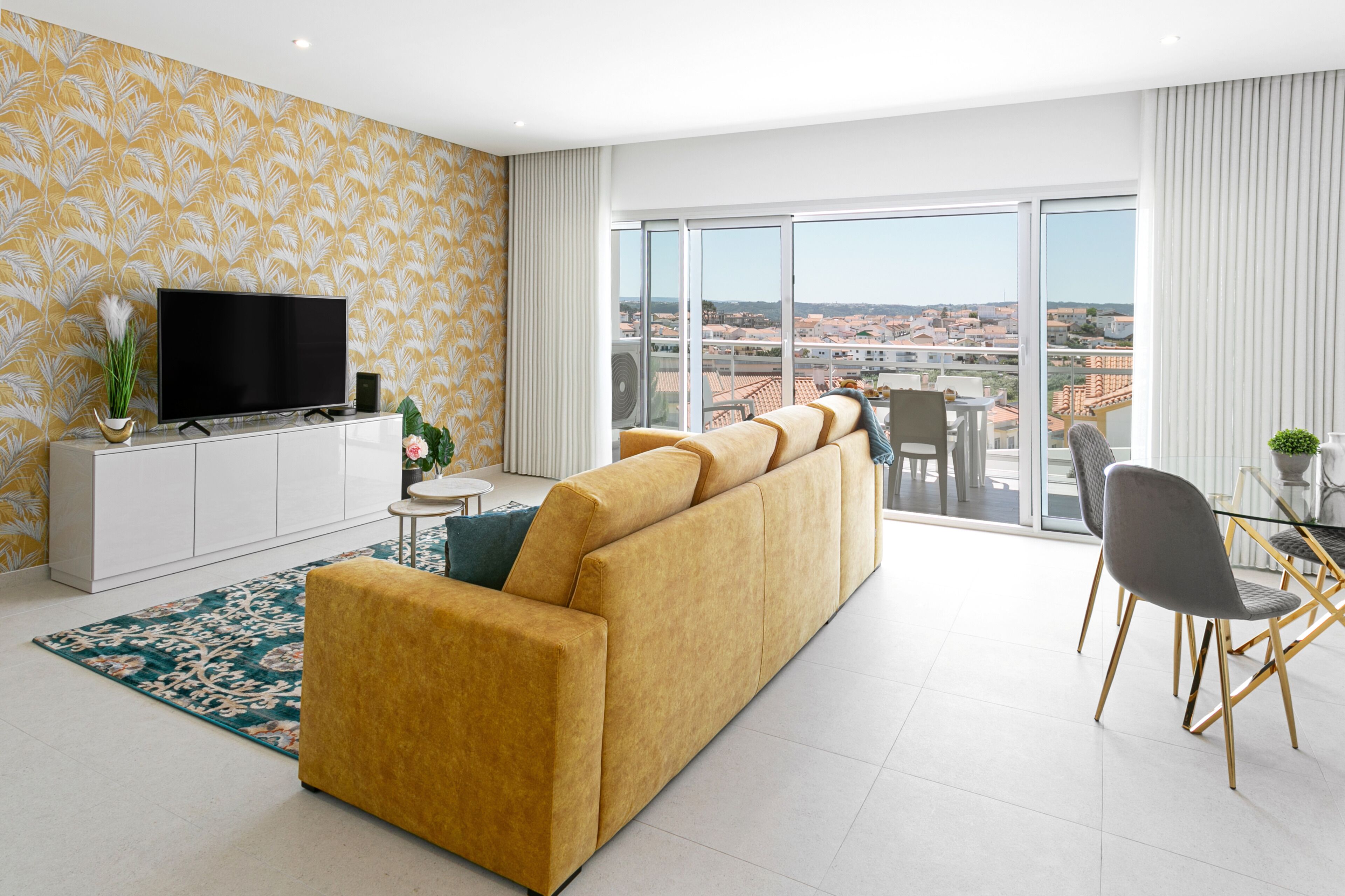 A modern living room with a sofa, TV, and dining area, featuring a city view through large windows.