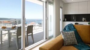 Un soggiorno moderno con un divano, una zona pranzo con tavolo e sedie, e un'ampia porta scorrevole in vetro che si apre su un balcone con vista sul mare.