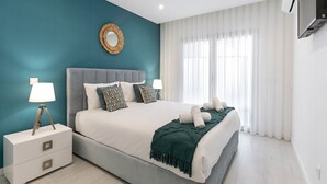 Una camera da letto moderna con una parete verde acqua, uno specchio rotondo, un letto imbottito grigio e un comodino bianco con una lampada.