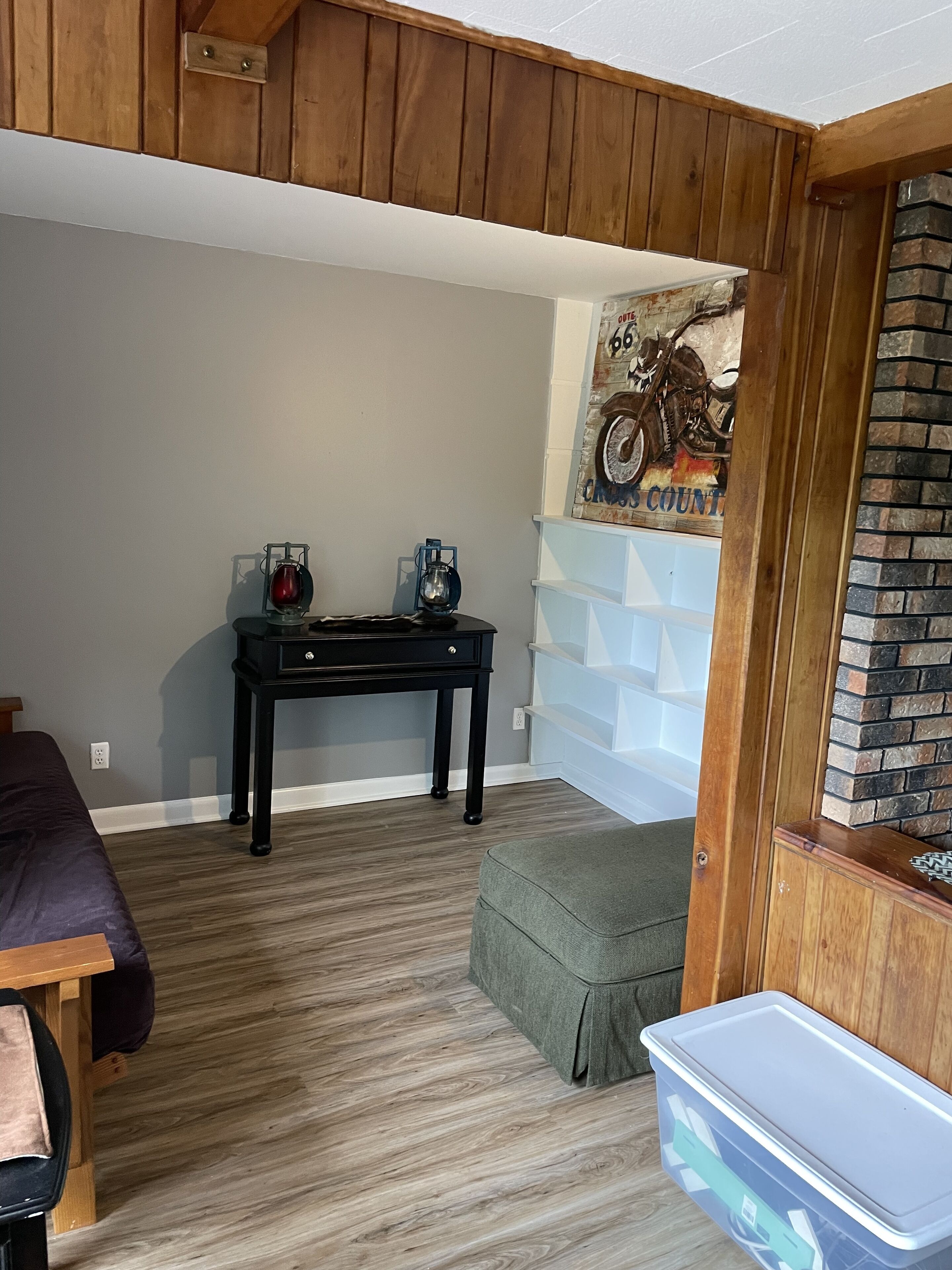 A room with a wooden ceiling, a motorcycle-themed wall art, a black table with two lanterns, a green ottoman, and a white storage bin.