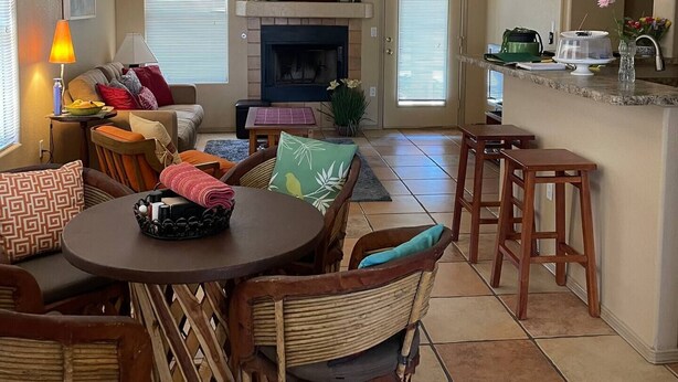 Un salon spacieux avec une cheminée, un coin repas avec des chaises en osier et une cuisine équipée de tabourets de bar.