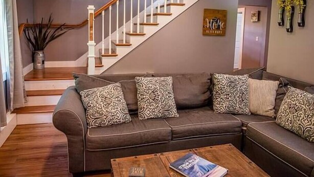 Un salon avec un canapé d’angle marron, des coussins décoratifs à motifs, une table basse en bois et un escalier avec des rampes blanches.