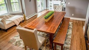 Un coin repas avec une table en bois, des bancs et un tapis. On aperçoit un canapé et un fauteuil au fond.