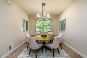 Une salle à manger avec une table ronde, six chaises, un lustre et une vue sur l’extérieur à travers une fenêtre munie de stores.