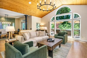 Un salon spacieux avec un plafond haut, des poutres apparentes, une grande fenêtre en arc de cercle et un coin salon confortable avec un canapé et des fauteuils.