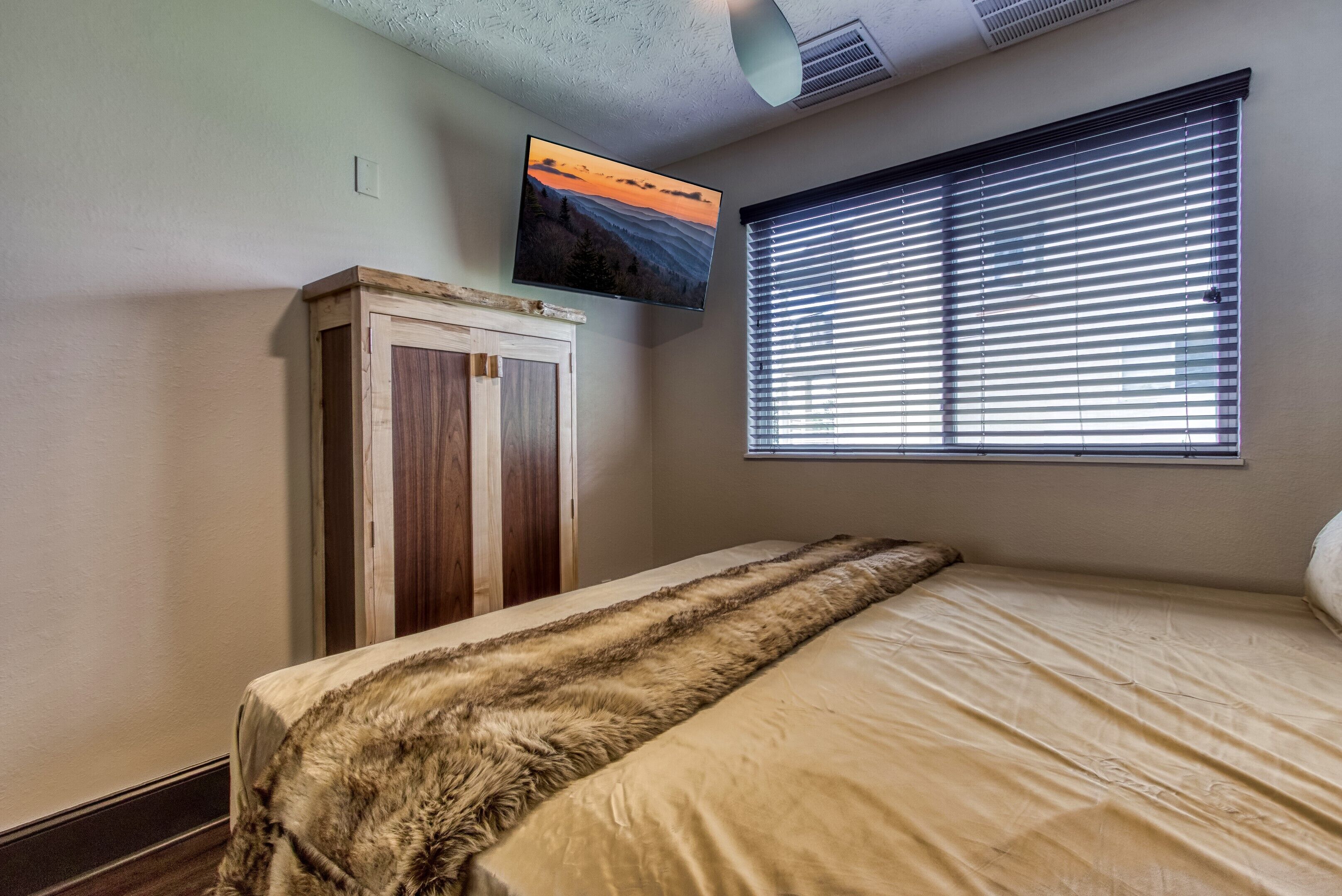 Une chambre à coucher avec un lit, une armoire en bois, une fenêtre avec des stores et un téléviseur fixé au mur.