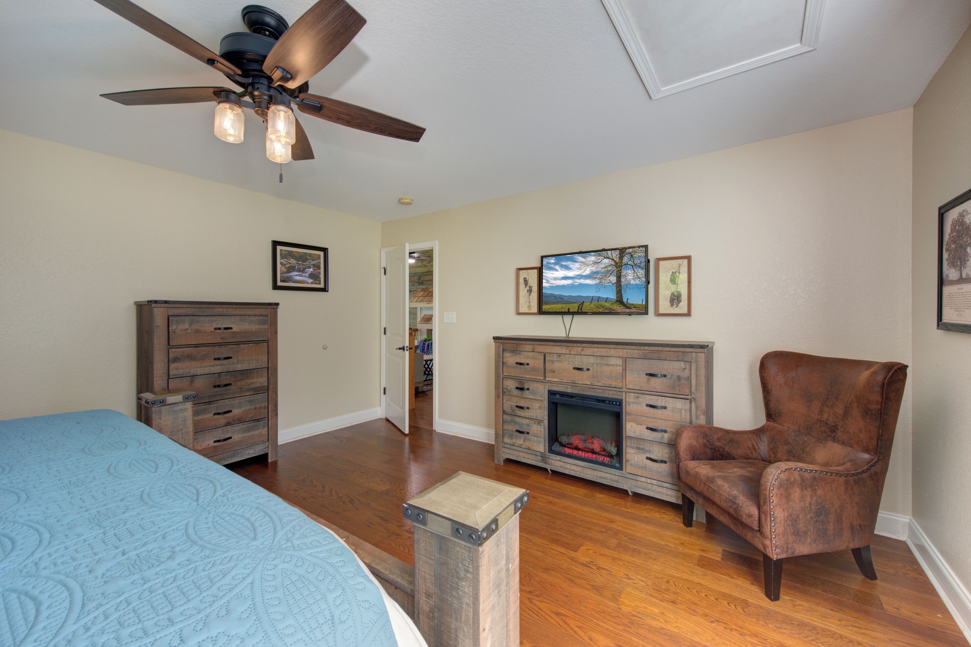 A bedroom with a bed, a wooden dresser, a leather armchair, a fireplace, and a ceiling fan.