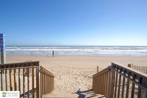 Une plage avec des passerelles en bois menant à l’eau, un ciel dégagé et une personne qui marche au loin.