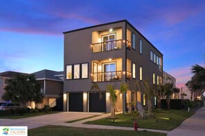 A modern two-story residential building with a balcony, large windows, and a well-lit driveway.