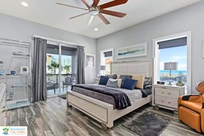Ein modernes Schlafzimmer mit Bett, Deckenventilator, Nachttisch und Blick auf den Strand durch ein großes Fenster.