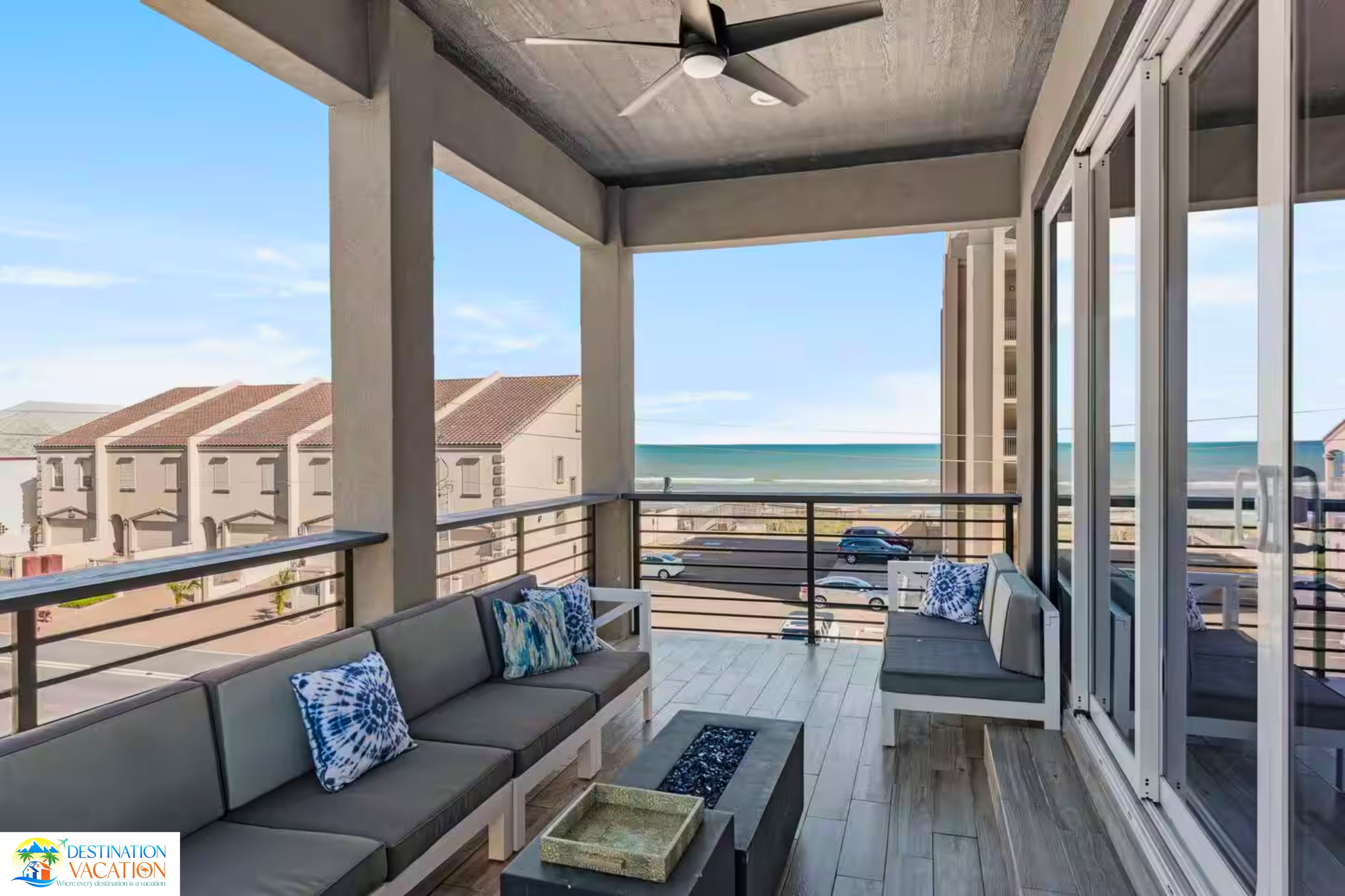 A balcony with a sofa and a ceiling fan, overlooking a beach and residential buildings.