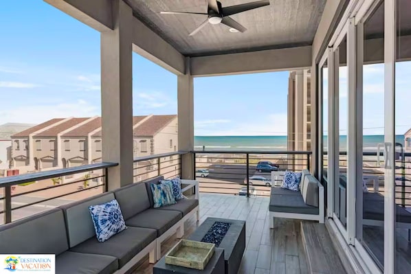 A balcony with a sofa and a ceiling fan, overlooking a beach and residential buildings.