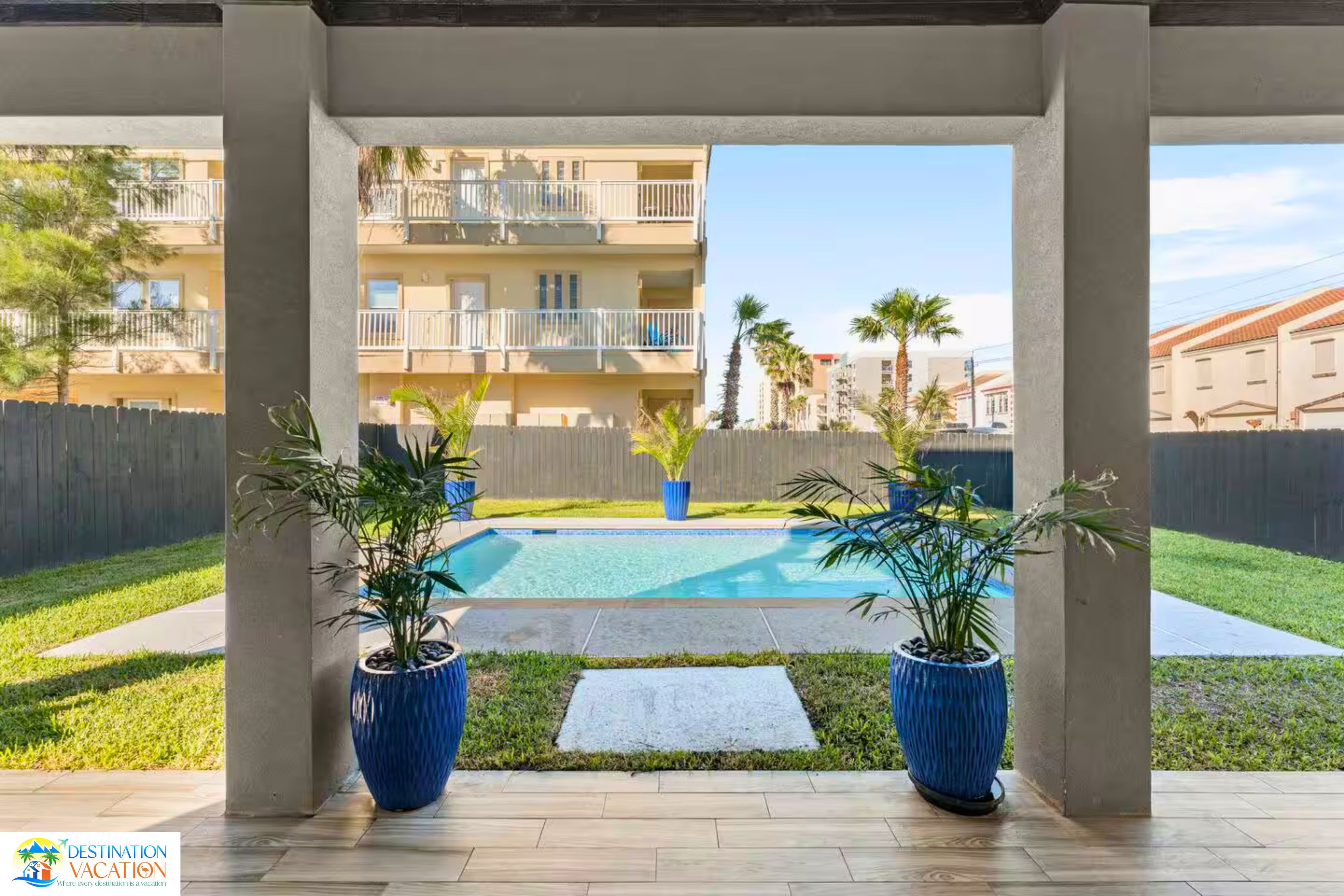 A pool area with two potted plants, a wooden fence, and a view of a multi-story building in the background.