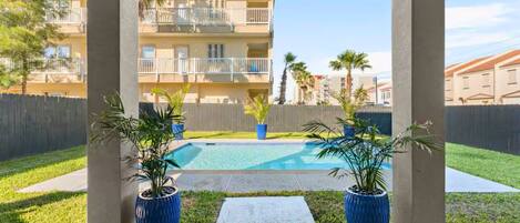 A pool area with two potted plants, a wooden fence, and a view of a multi-story building in the background.