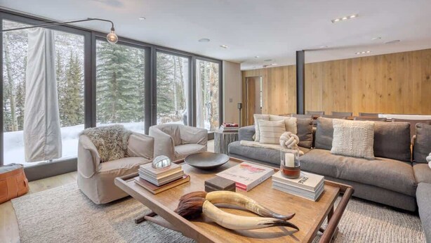 A modern living room with large windows, a sofa set, a coffee table with books and decorative items, and a wooden accent wall. - Luxury Ski-In/Out Residence at Golden Peak (Vail)